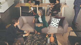 A number of people in a co-working space. Two people at a desk, and two other people on couches.