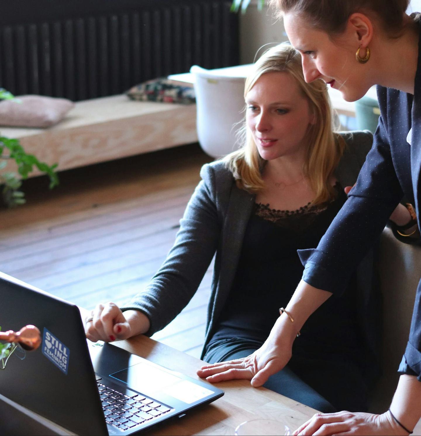 Two women seated at a desk.  They are looking at a laptop screen.
