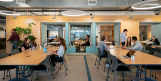 A vibrant coworking space with modern amenities featuring spacious communal tables, cozy booths, and abundant natural light. The area is bustling with professionals collaborating, working on laptops, and engaging in discussions. Contemporary lighting and greenery enhance the inviting atmosphere, making it an ideal environment for productivity and networking.