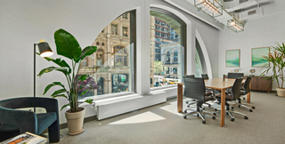A bright, modern office space featuring large arched windows with a view of historic buildings in New York City. The room includes a wooden conference table with black office chairs, contemporary lighting fixtures, and lush indoor plants. Comfortable seating and minimalist decor create an inviting and professional atmosphere.