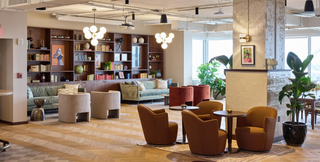 A sophisticated coworking lounge featuring plush seating, elegant lighting fixtures, and a wall of bookshelves filled with books and decorative items. The space includes comfortable sofas, armchairs, and small round tables, creating an inviting atmosphere for work or relaxation. Large windows and indoor plants enhance the bright and airy feel of the room.