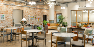 A chic and inviting coworking space featuring exposed brick walls, modern lighting fixtures, and a variety of seating options. The area includes round marble tables, cozy armchairs, and sofas, creating a comfortable and stylish environment. Indoor plants and eclectic decor add warmth and character to the space.