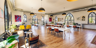 A spacious, modern coworking area featuring large arched windows, natural light, and contemporary furniture. The room has a mix of workstations and comfortable seating areas with vibrant decor and indoor plants, creating a stylish and productive environment.