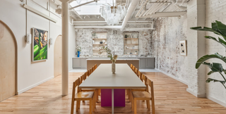 A modern, loft-style dining space featuring a long table with wooden chairs, situated under a chandelier with circular light fixtures. The room boasts a white, exposed brick wall, wooden floors, and a combination of open shelving and cabinetry along the back wall. Artwork adorns the walls, adding a touch of sophistication. The space is bathed in natural light from large windows or skylights above, and a potted plant adds a hint of greenery to the right side.